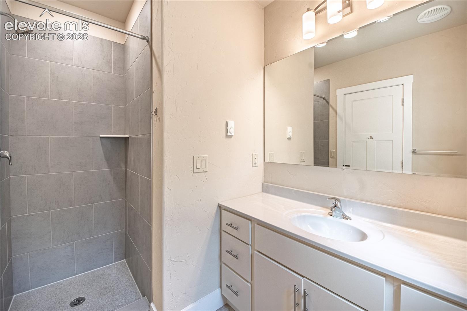 Bathroom featuring a walk-in shower with large format grey tile, a built-in shower shelf, a vanity with white cabinetry, a white countertop, and a wide mirror with overhead lighting