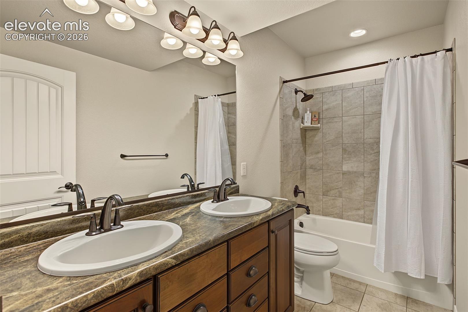 Dual-sink vanity with stone-finish countertop, dark wood cabinetry, multi-bulb vanity lighting, and shower-tub combination with tile surround