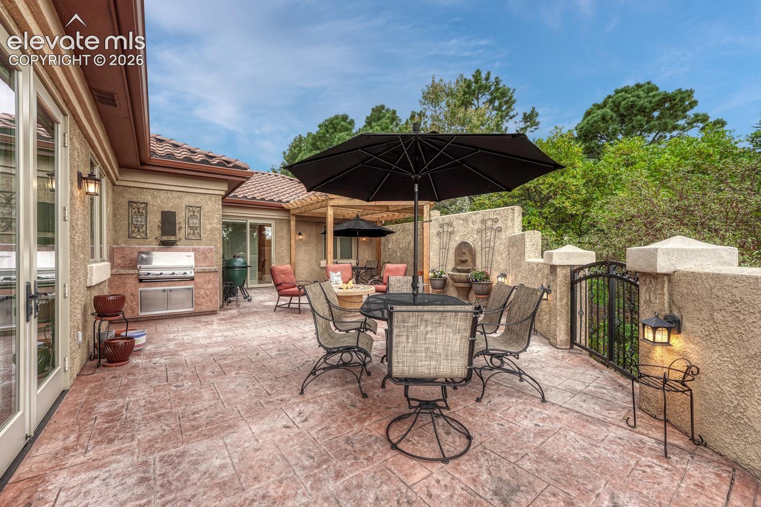 Stamped concrete patio featuring a built-in outdoor stainless steel grill, wooden pergola, decorative wall-mounted fountain, and perimeter stucco wall with iron gate