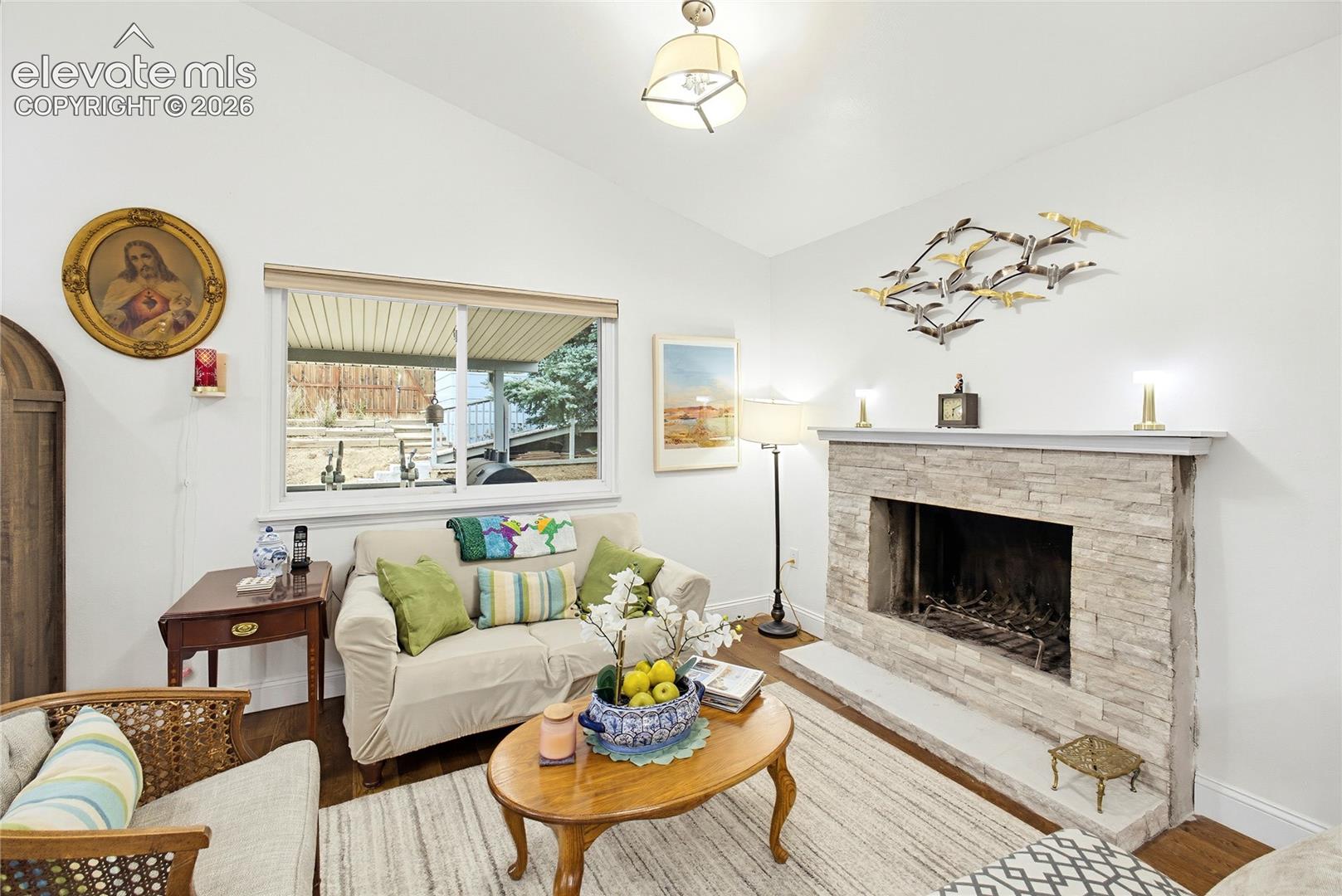 Vaulted ceiling, stone-clad fireplace, wood-finish flooring, built-in fireplace grate, and a large window