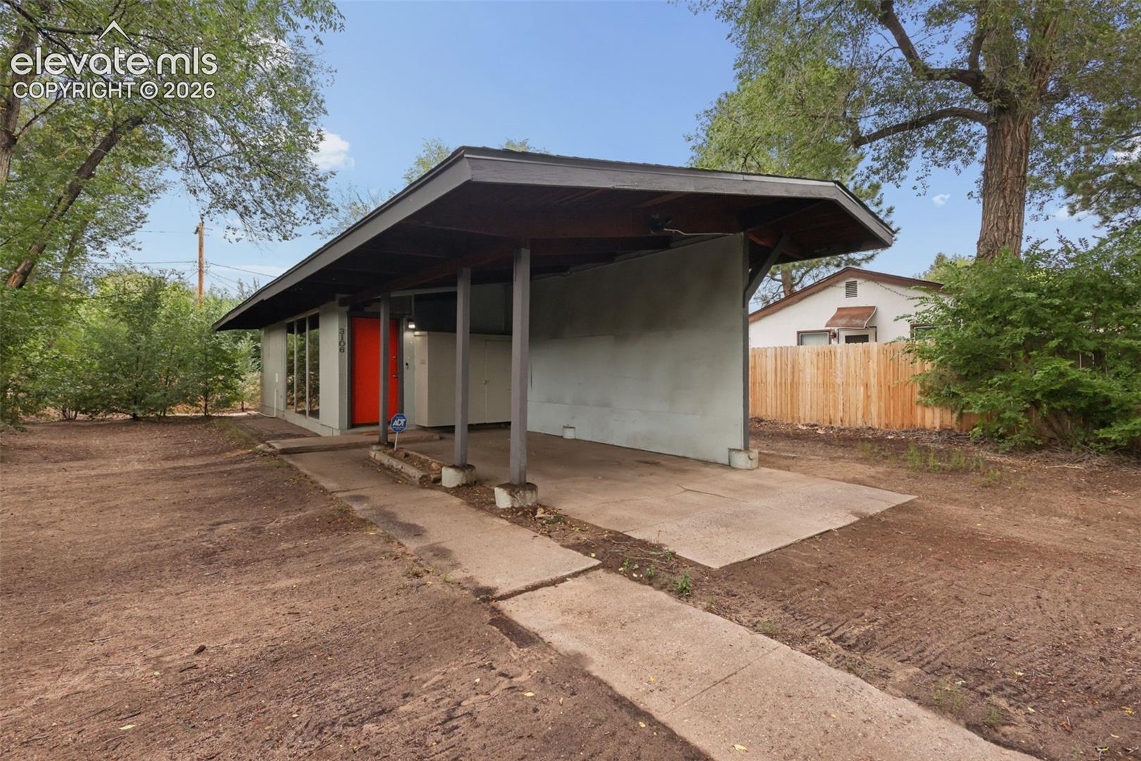Exterior featuring a covered carport, red entry door, horizontal window bank, and mature perimeter trees