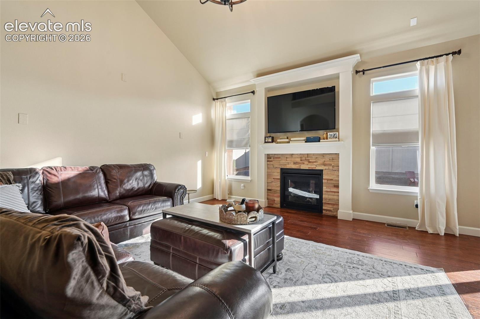 Vaulted ceiling with a central lighting fixture, hardwood-finish flooring, stone-faced fireplace, white trim, and multiple windows