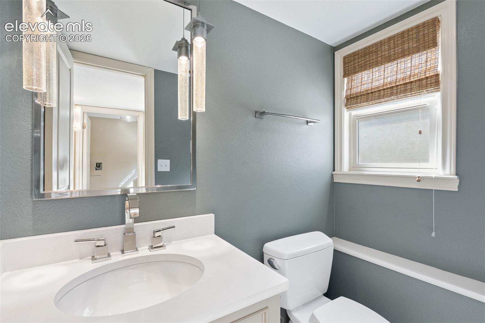 Bathroom featuring a white vanity with integrated basin, polished metal hardware, rectangular wall-mounted mirror, cylindrical pendant lighting, and a window with custom shade