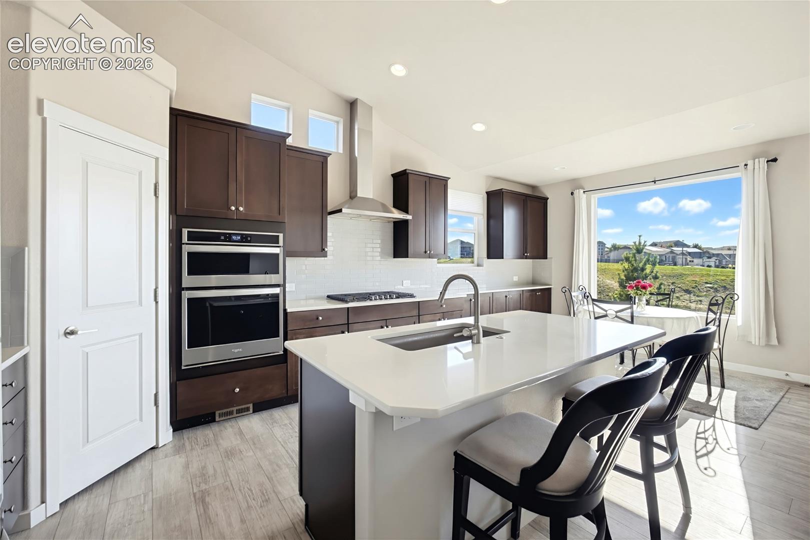 Kitchen featuring a central island with a quartz countertop, dark wood-finish cabinetry, stainless steel double wall ovens, a gas cooktop with a chimney-style range hood, and tile flooring