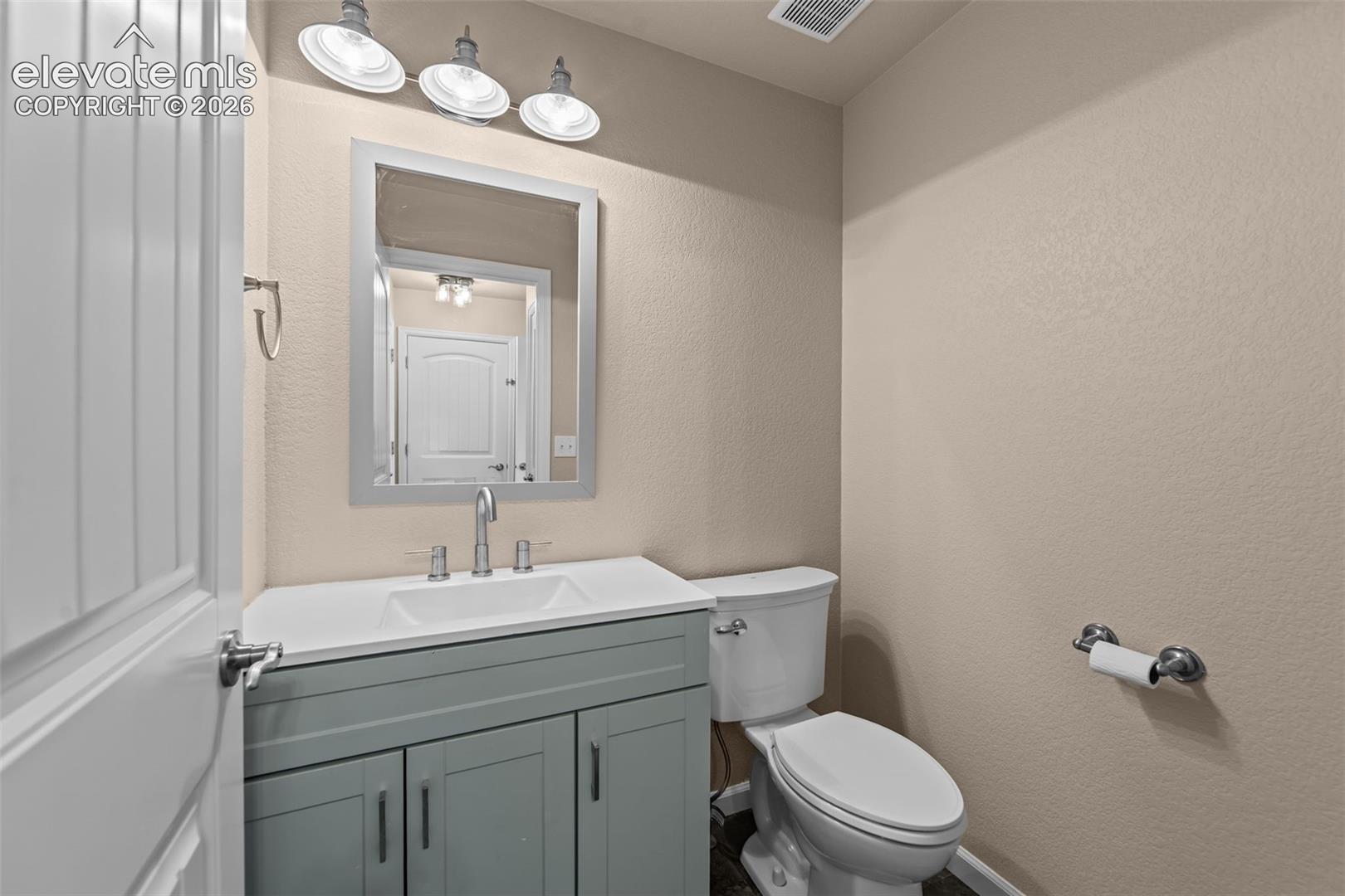 Vanity featuring a solid surface countertop with integrated basin, blue-gray cabinetry with metallic hardware, three-light industrial-style wall fixture, rectangular framed mirror, and chrome faucet