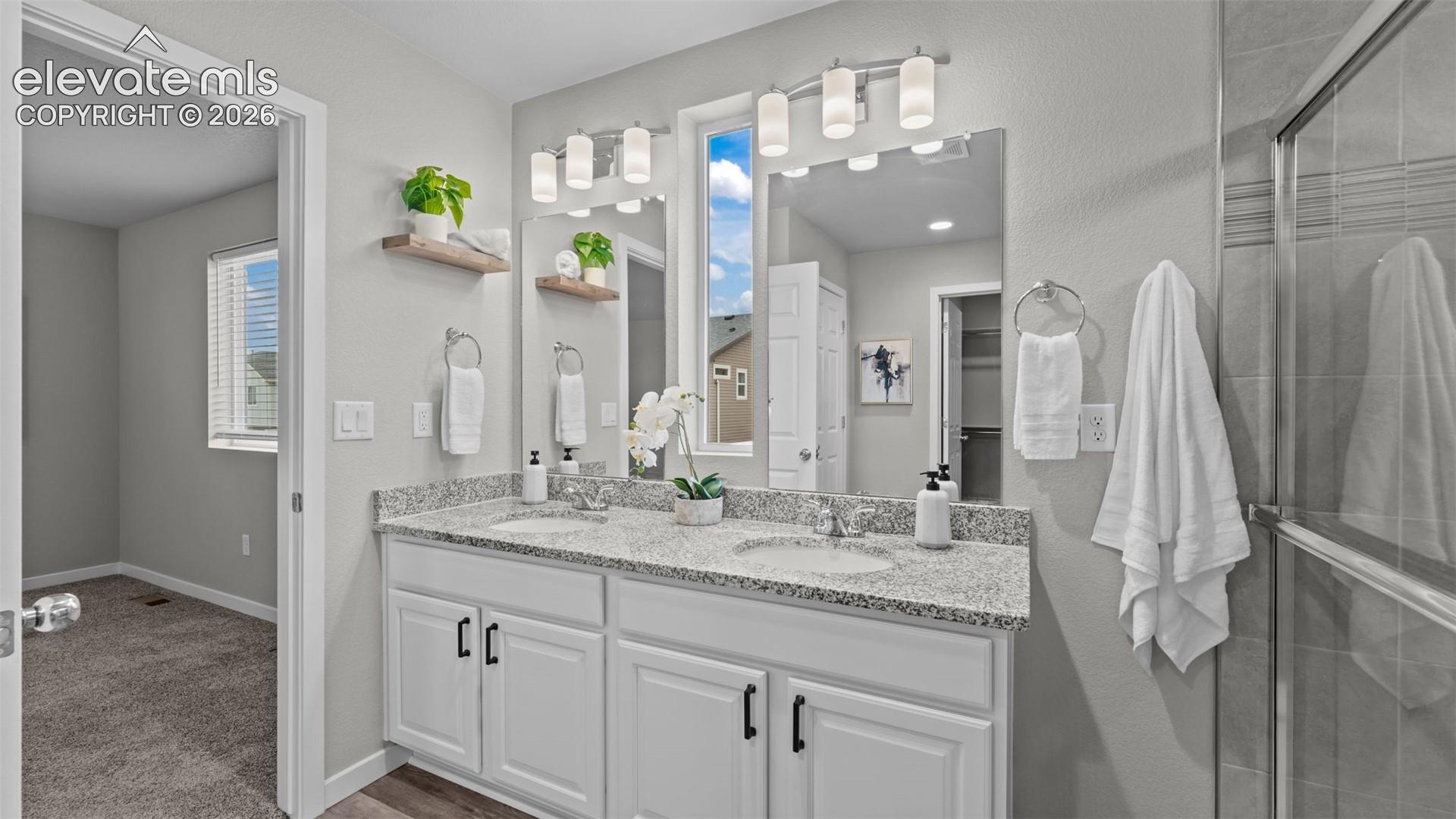 Dual-sink vanity with stone-finish countertop, white shaker-style cabinetry featuring matte black hardware, dual wall-mounted vanity lights, and a glass-enclosed shower
