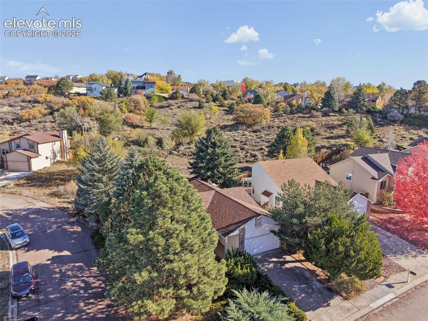 Aerial perspective of residential property featuring an attached garage, paved driveway, expansive sloping terrain, and mature evergreen trees