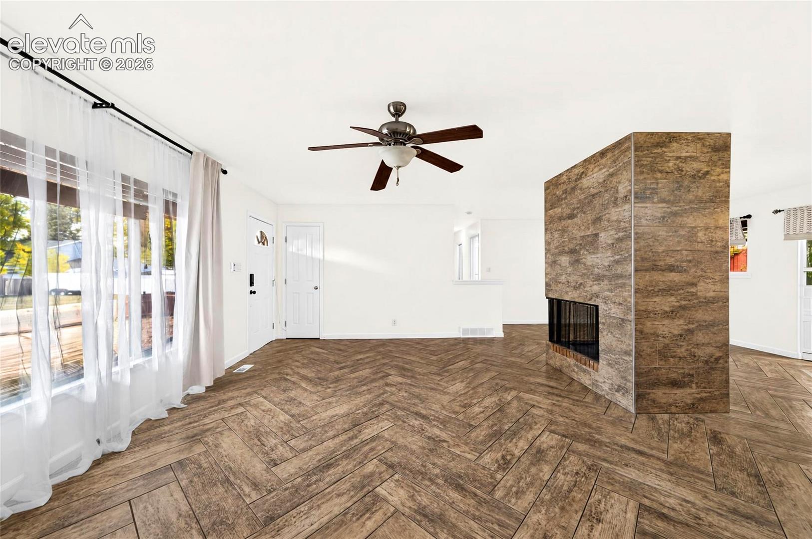 Herringbone-patterned wood-finish flooring, central fireplace with floor-to-ceiling wood-finish tile surround, large picture windows, and a multi-blade ceiling fan