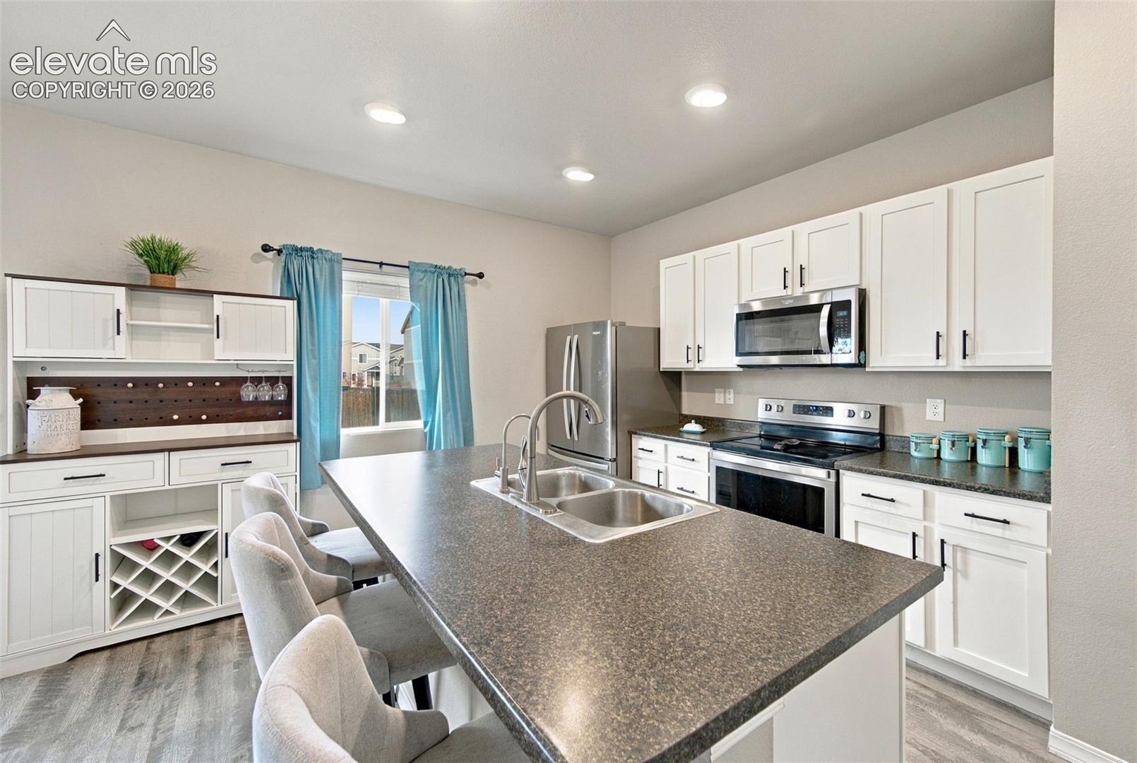 Kitchen featuring white shaker cabinetry, an expansive center island with a dual-basin stainless steel sink, dark solid-surface countertops, stainless steel appliances, and built-in hutch cabinetry with integrated wine storage