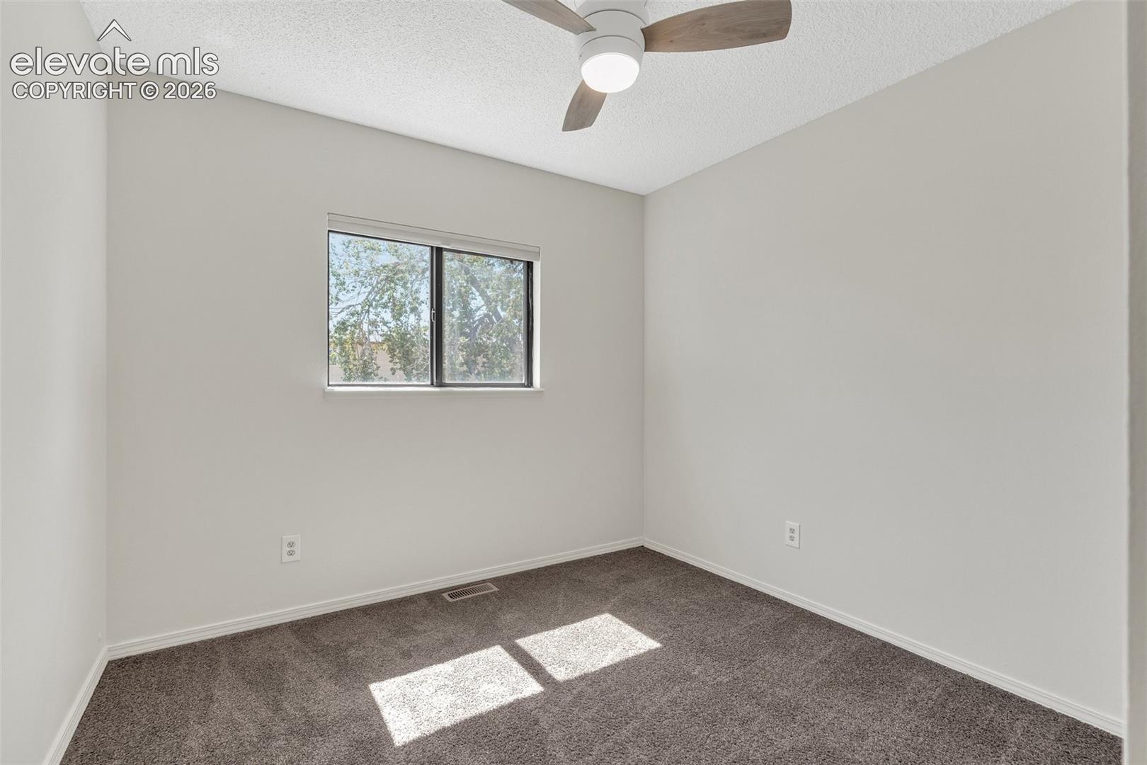 Light-colored walls, window with natural light, dark carpeting, and a ceiling fan with integrated lighting