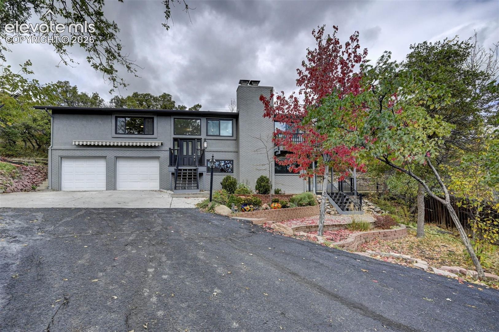 Split-level exterior featuring a painted brick facade, two-car garage with white paneled doors, tiered stone retaining walls, and metal stair railings