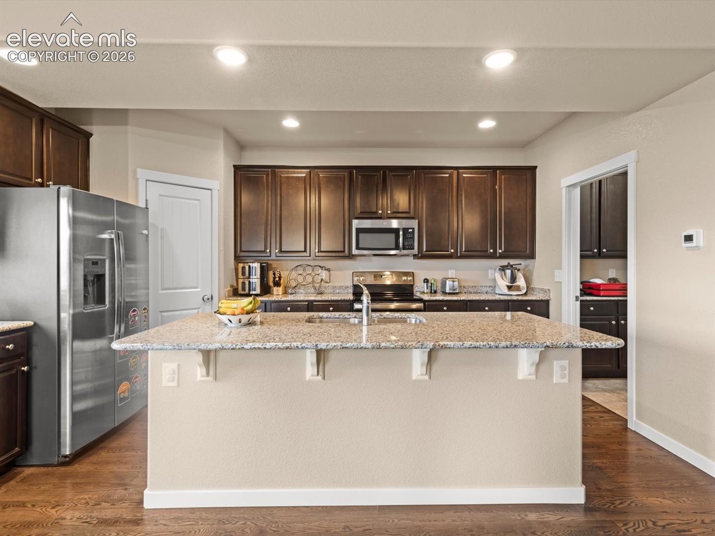 Central kitchen island with a speckled countertop, dark wood-finish cabinetry, stainless steel appliances, and recessed lighting