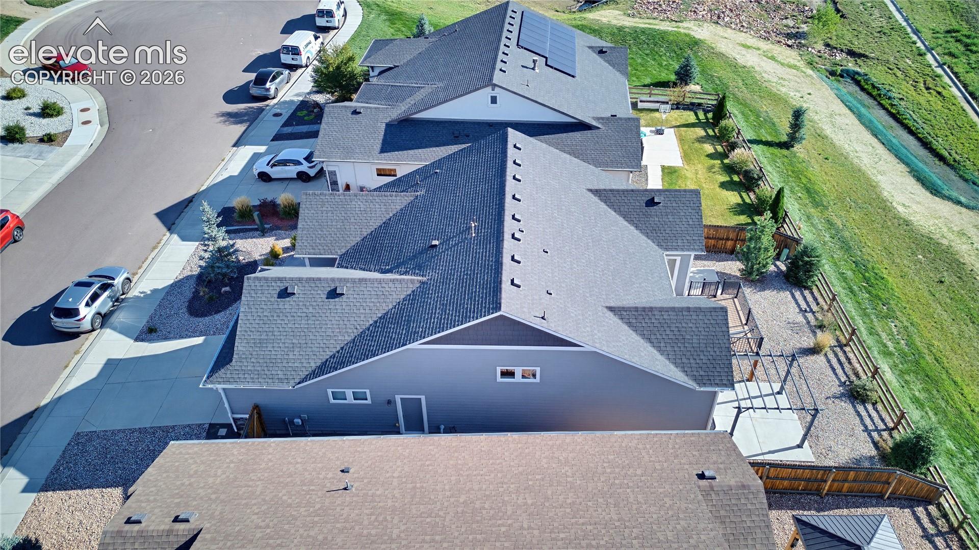 Aerial view of a residential property featuring a gray exterior, asphalt shingle roofing, integrated solar panels, a concrete patio, and a pergola
