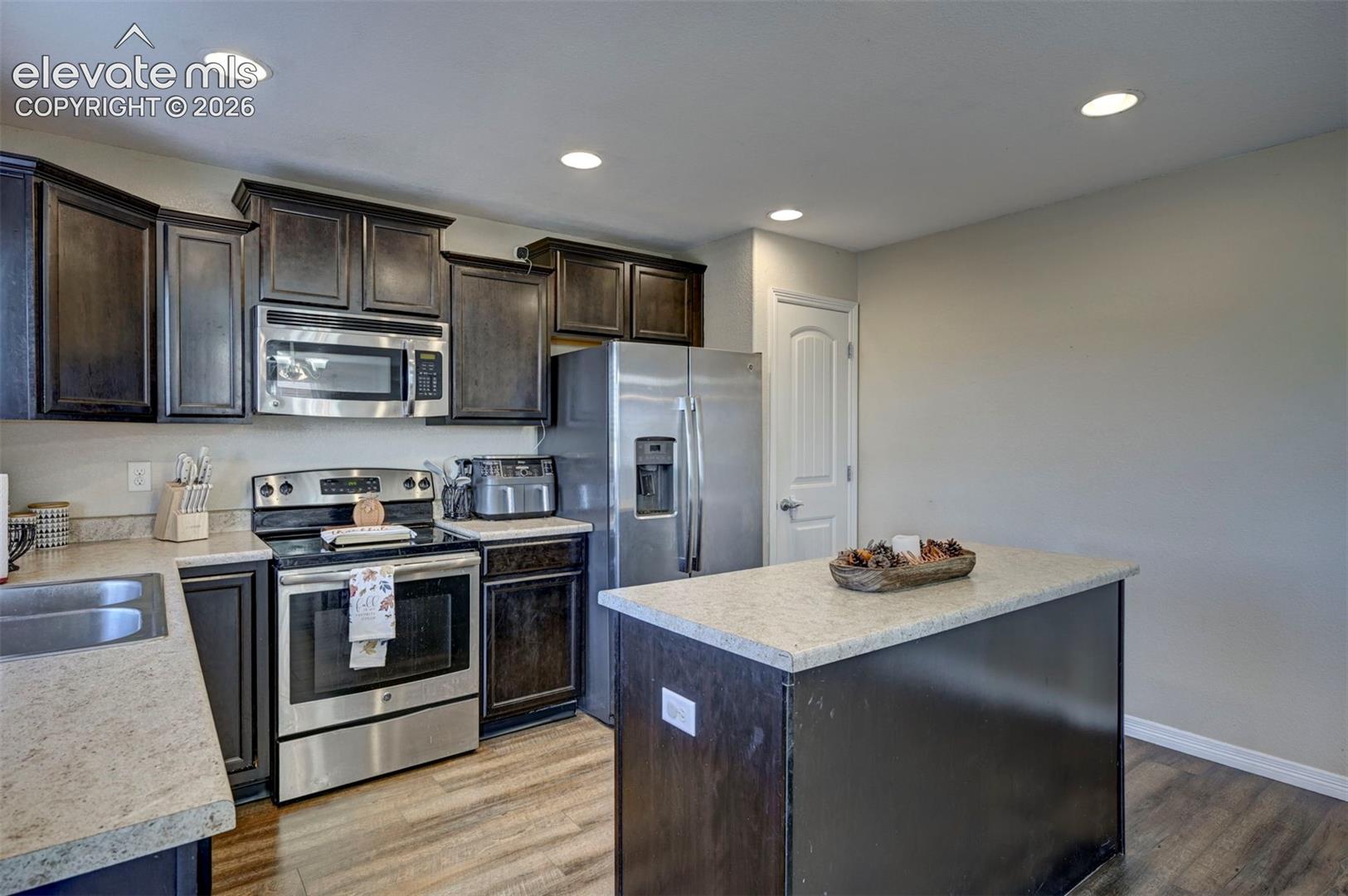 Kitchen featuring dark wood-finish cabinetry, stainless steel appliances, light-toned countertops, and wood-finish flooring
