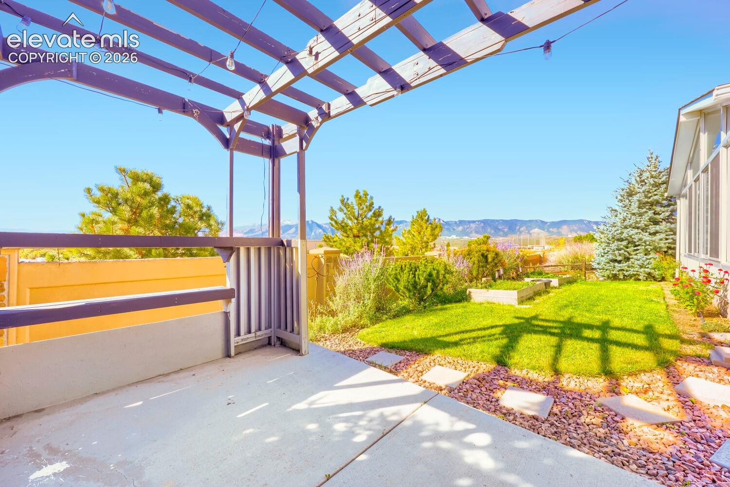 Concrete patio featuring an overhead wooden pergola, landscaped lawn, stone paver accents, and decorative rock beds against a mountain backdrop