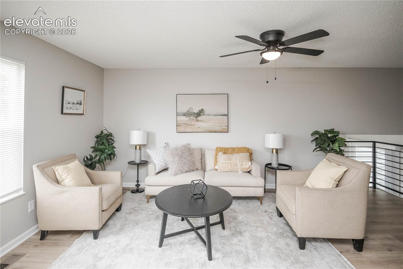 Living area featuring wood-finish flooring, a matte black ceiling fan with integrated lighting, and a metal horizontal-bar railing