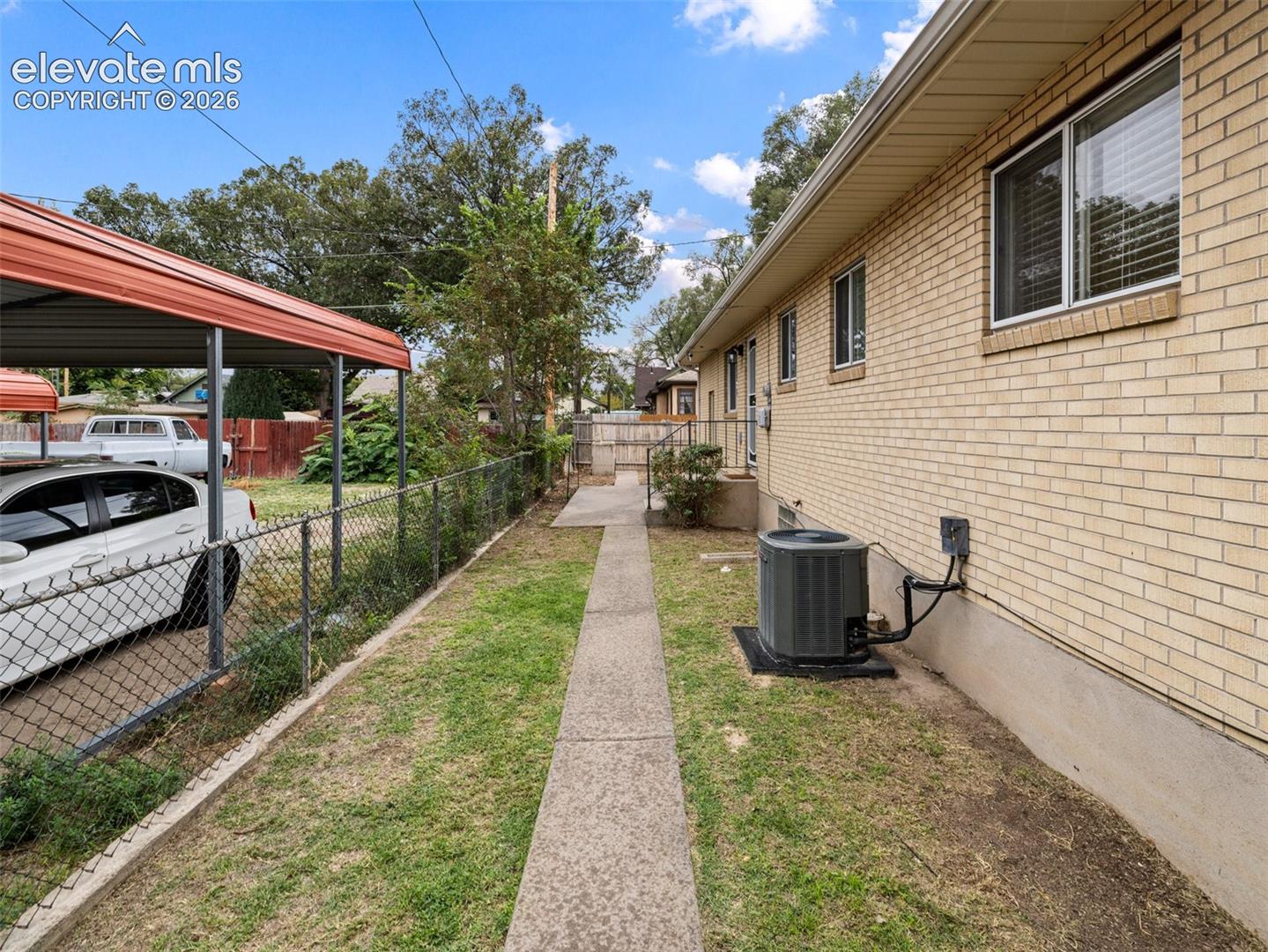 Brick exterior with a concrete walkway, chain link fencing, and a red metal carport