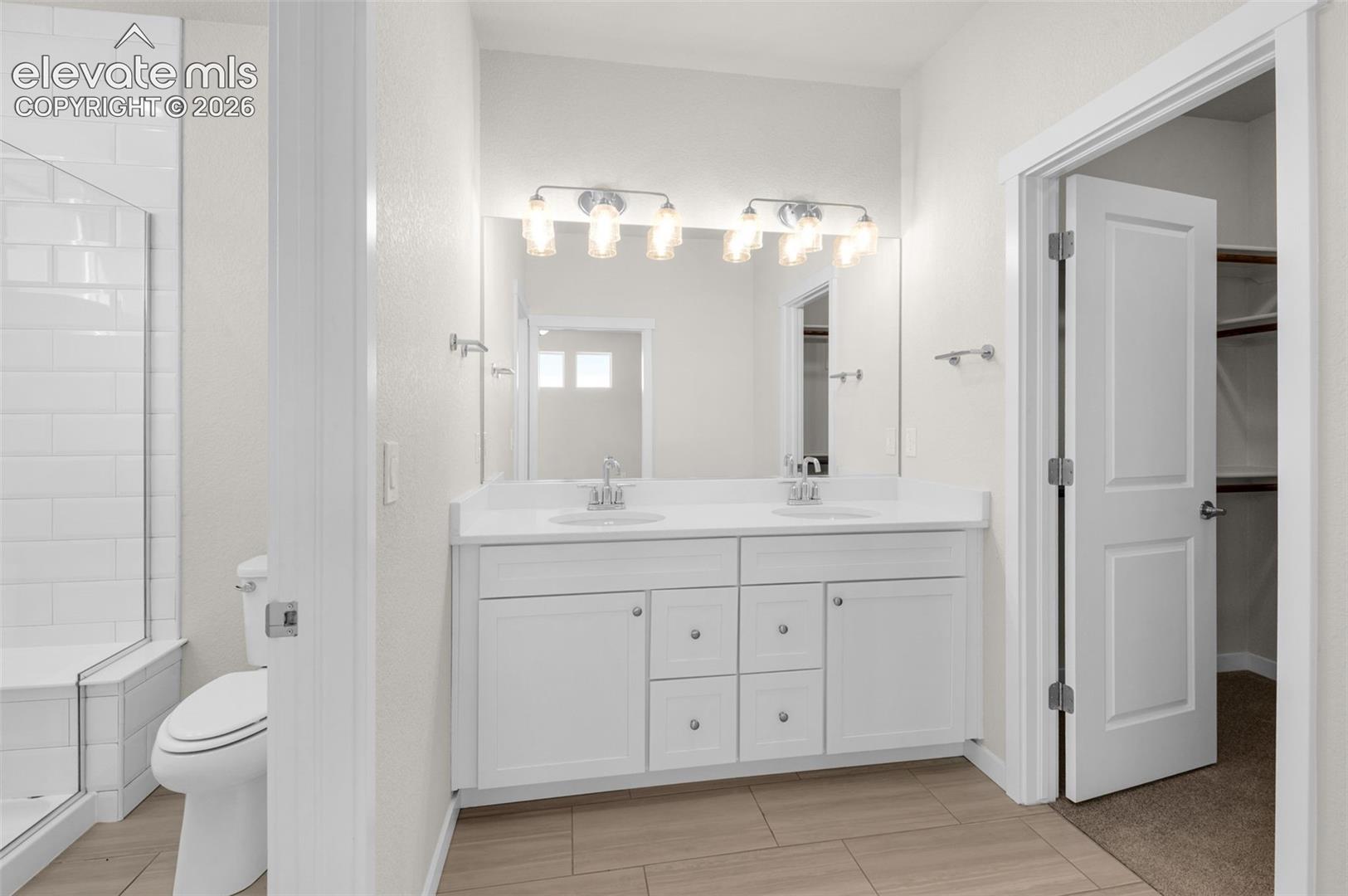 Dual-sink vanity with white cabinetry and solid-surface countertop, full-width vanity mirror, polished chrome lighting fixtures, and walk-in shower with white subway tile surround