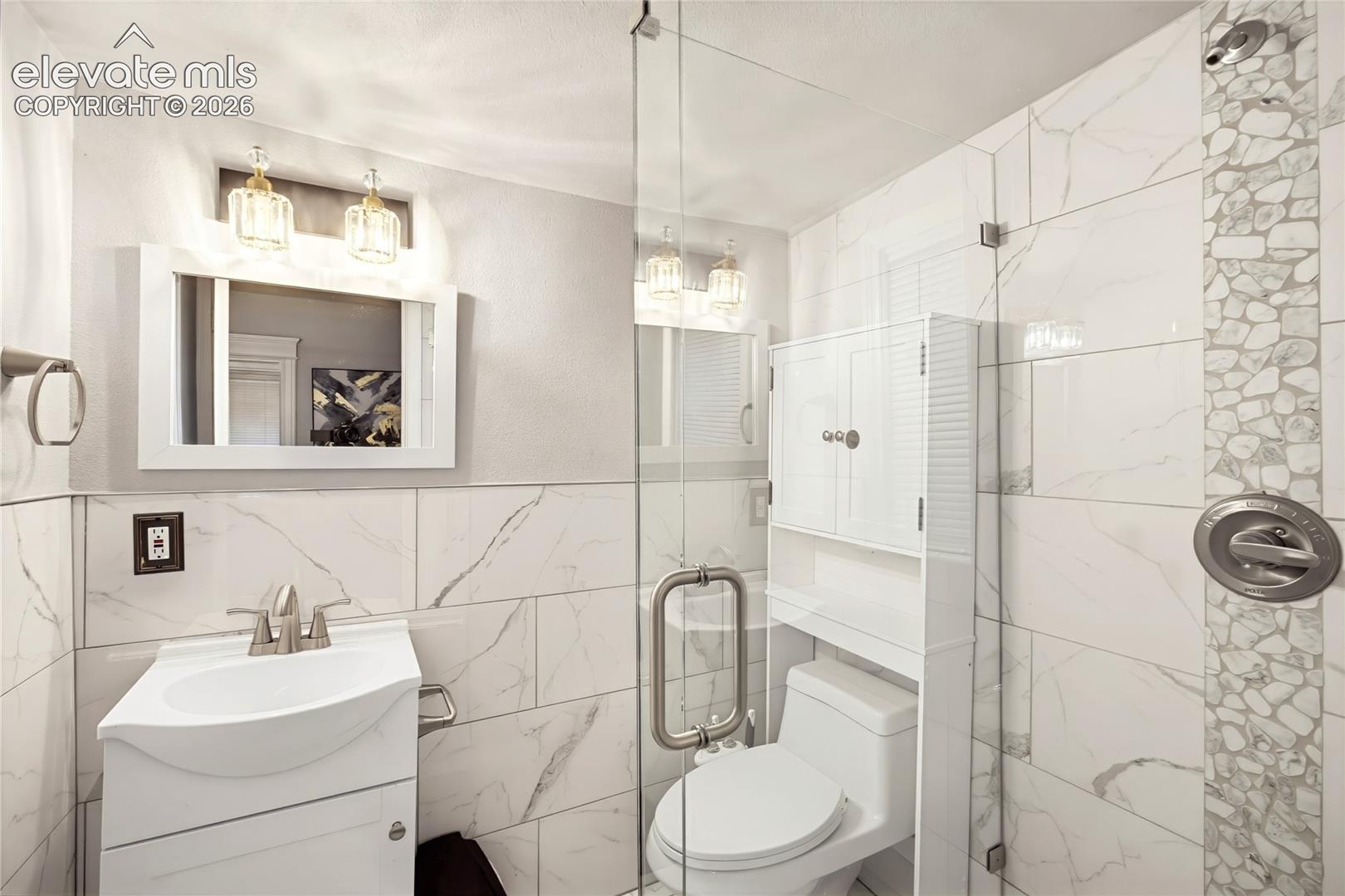 Bathroom featuring a walk-in shower with clear glass enclosure, white rectangular vanity with integrated sink, sleek vanity light fixtures, and white tiled walls