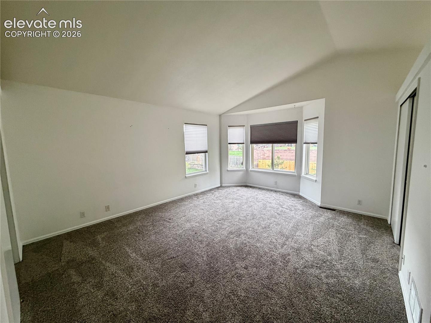 Spacious room featuring vaulted ceilings, a bay window with multiple panes, gray carpet flooring, white walls, and a white bi-fold closet door