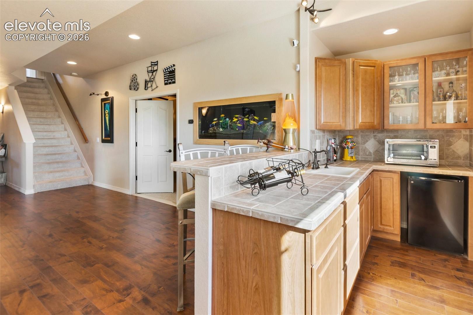 Kitchen area featuring wood-finish flooring, tile-surround bar top, wood cabinetry with glass-front display panels, recessed lighting, and a built-in wall aquarium