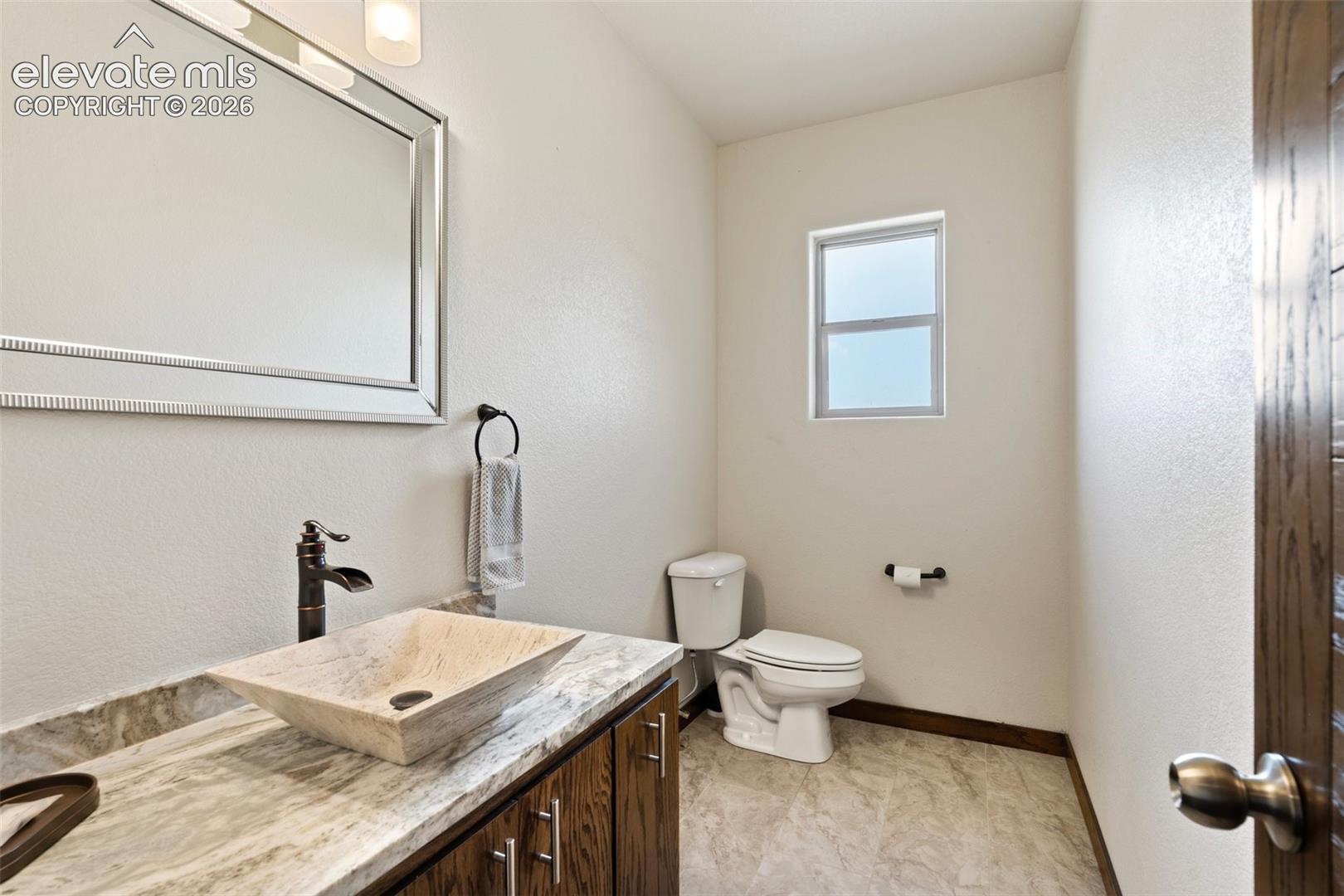 Bathroom featuring a vessel sink with a dark bronze faucet, a vanity with wood-finish cabinetry and stone-look countertop, a framed mirror, and a frosted window