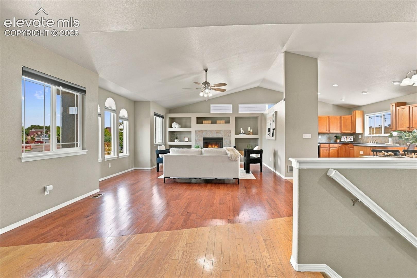 Vaulted ceilings, wood-finish flooring, built-in shelving, stone-surround fireplace, and a ceiling fan with integrated lighting