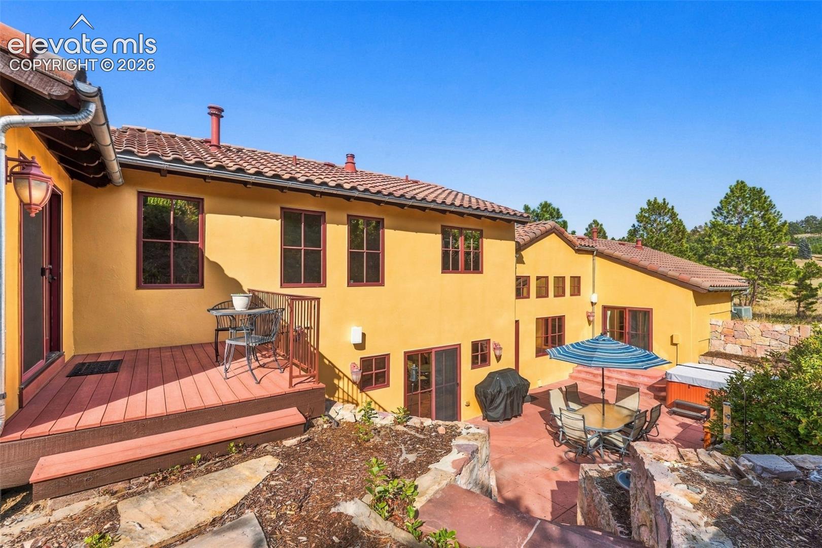 Stucco exterior with a tile roof, an elevated wood-finish deck, and a stone patio featuring terraced landscaping