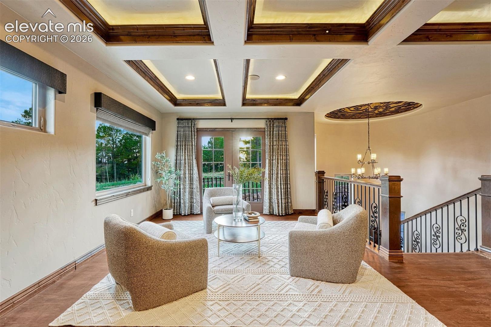 Coffered ceiling with integrated lighting, wood-finish flooring, French doors with glass panels, wrought iron stair railing, and a decorative ceiling medallion with a chandelier