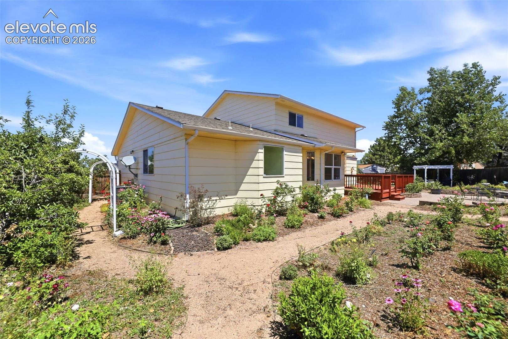 Exterior siding in a light hue, featuring a multi-level deck with red railing, a gravel pathway, and garden beds with plantings