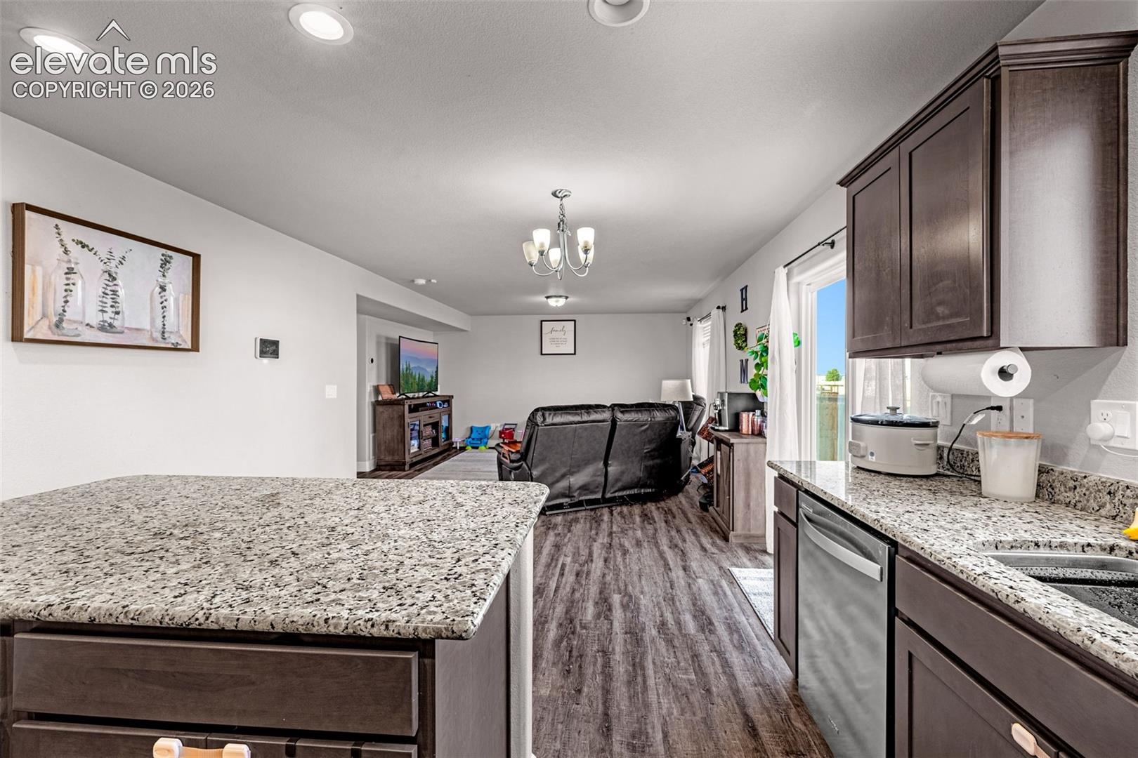 Open-concept living area featuring a kitchen island with granite-finish countertop, dark wood-finish cabinetry, stainless steel dishwasher, and wood-finish flooring extending into the living space