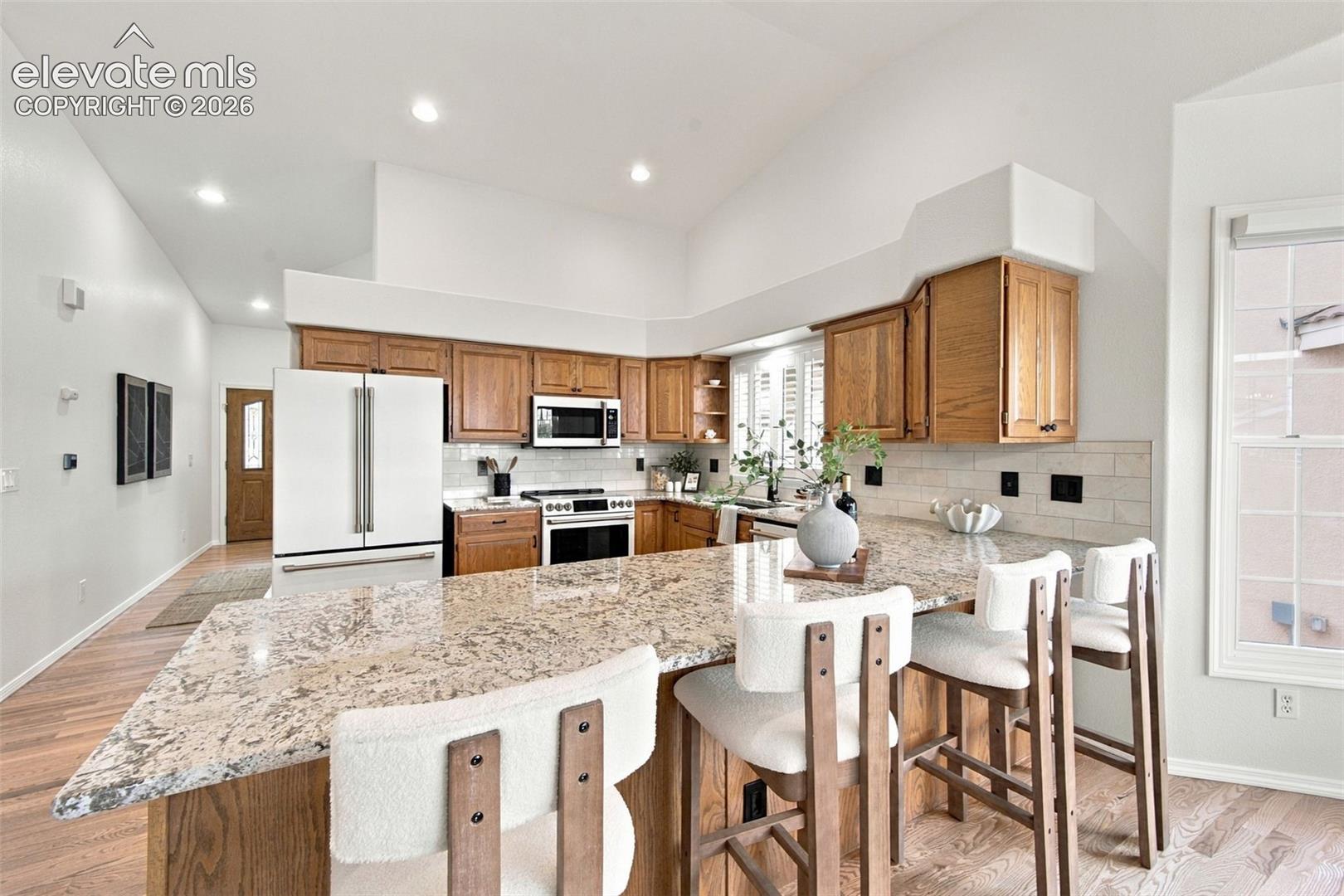 Kitchen featuring vaulted ceilings, stone-surface breakfast bar, wood cabinetry, light-tone tile backsplash, and wood-finish flooring