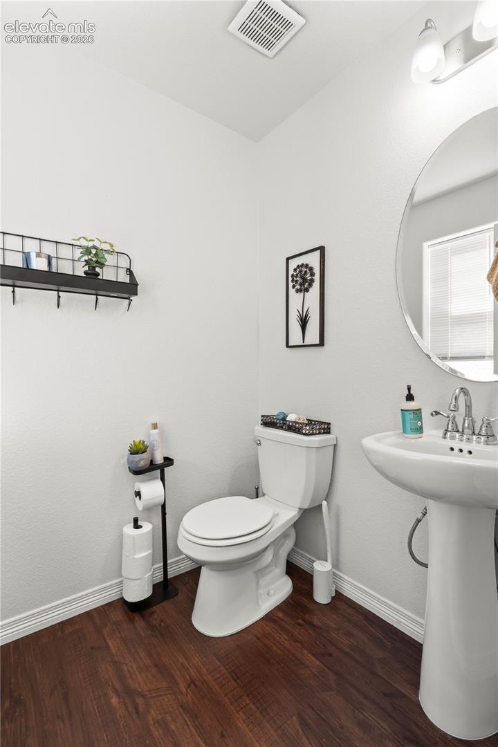 Pedestal sink with chrome faucet, circular wall-mounted mirror, white ceramic toilet, and wood-finish flooring