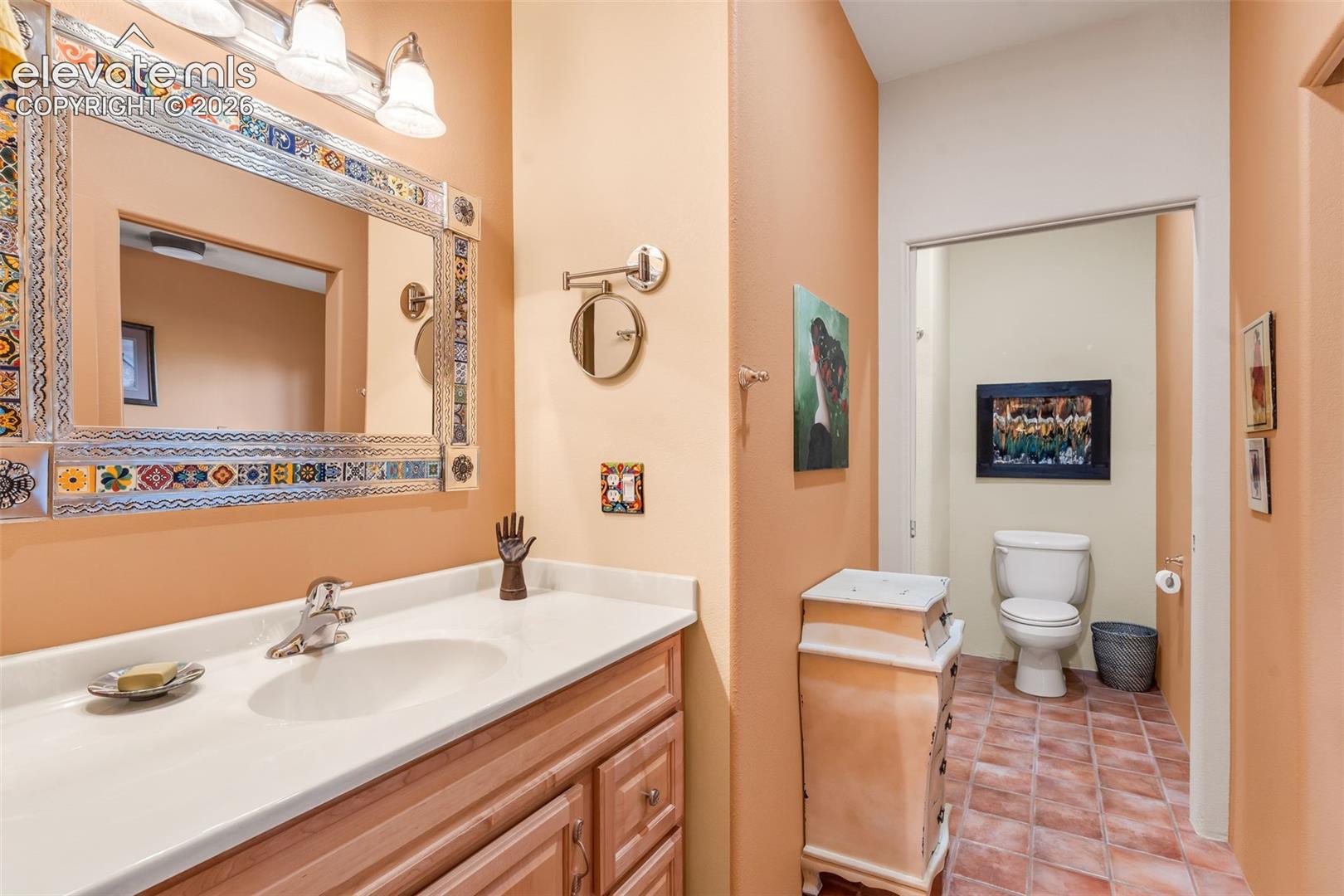 Vanity featuring light wood-finish cabinetry, white countertop with an integrated basin, metal-framed decorative mirror, three-light fixture, and tile flooring