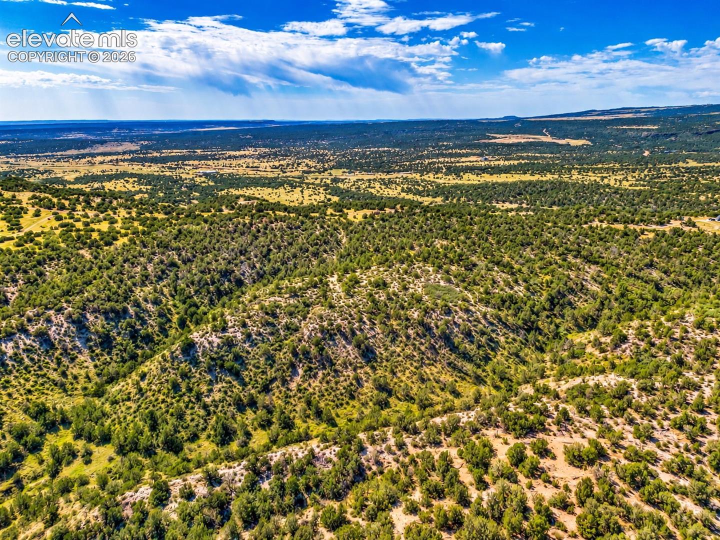 Scenic aerial perspective of rolling hills, natural rock outcroppings, and scattered juniper trees with beautiful mountain ranges in the distance.

