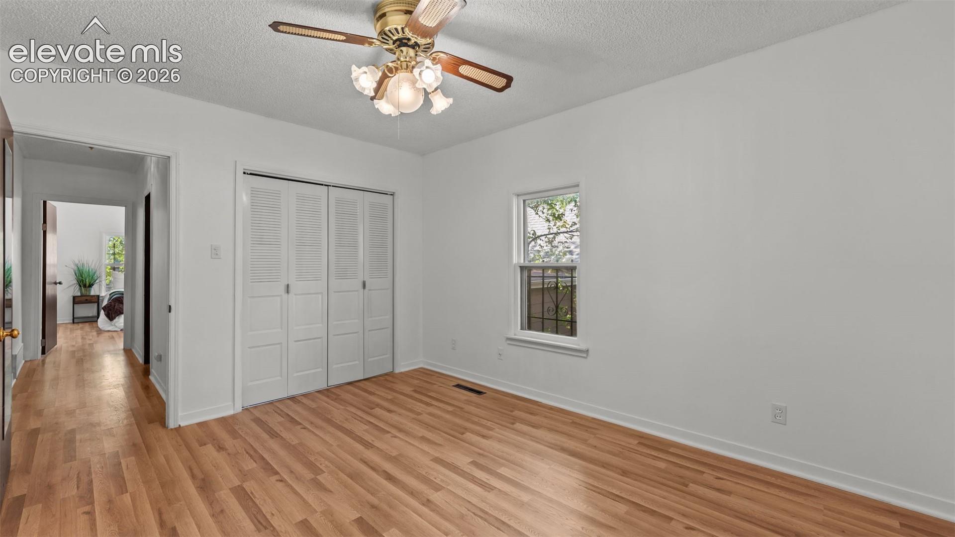 Spacious bedroom featuring warm wood-finish flooring, a ceiling fan with integrated lighting, large closet with bi-fold doors, and abundant natural light from a side window