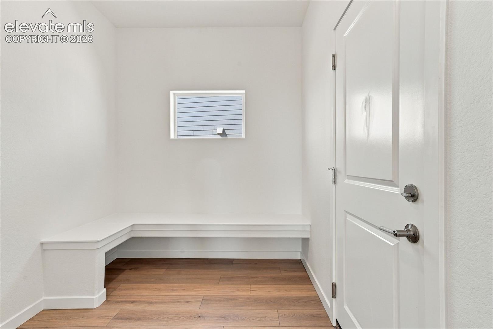 White paneled door with brushed nickel hardware, built-in white bench, wood-finish flooring, and a single window with blinds