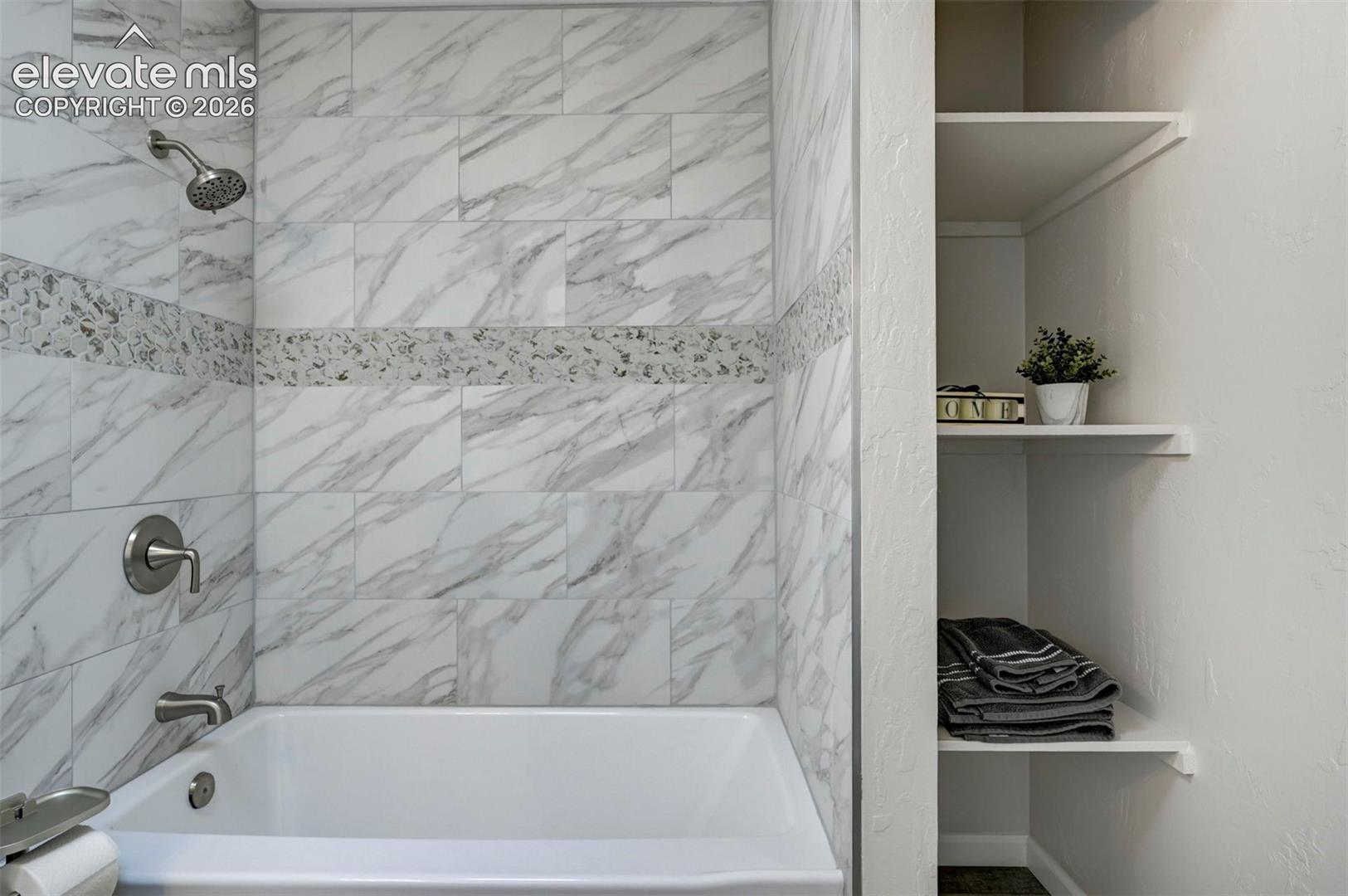 Shower-tub combination featuring large-format stone-patterned wall tile, a decorative mosaic accent band, brushed nickel fixtures, and built-in recessed shelving