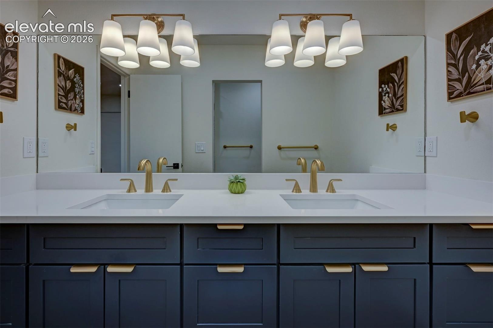 Dual-sink vanity with navy shaker-style cabinetry, white solid-surface countertop, gold-tone hardware, and dual four-light wall fixtures