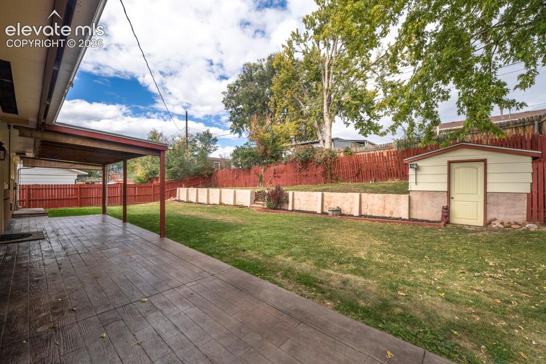 Covered patio featuring stamped concrete flooring, tiered stone retaining walls, enclosed yard with wood fencing, and a detached storage shed