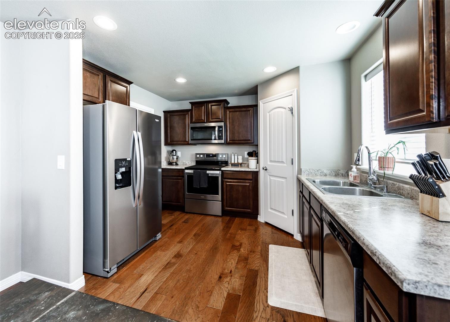 Kitchen featuring dark-finish wood cabinetry, stainless steel appliances, stone-patterned laminate countertops, double-basin stainless steel sink, and wood-finish flooring