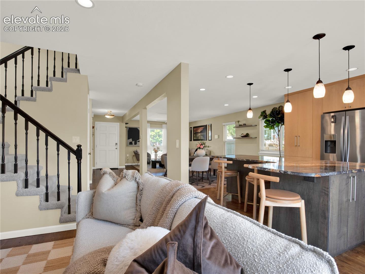 Open-concept interior featuring an island with stone-finish countertop, stainless steel refrigerator, recessed lighting, triple pendant light fixtures, and a staircase with a black metal railing