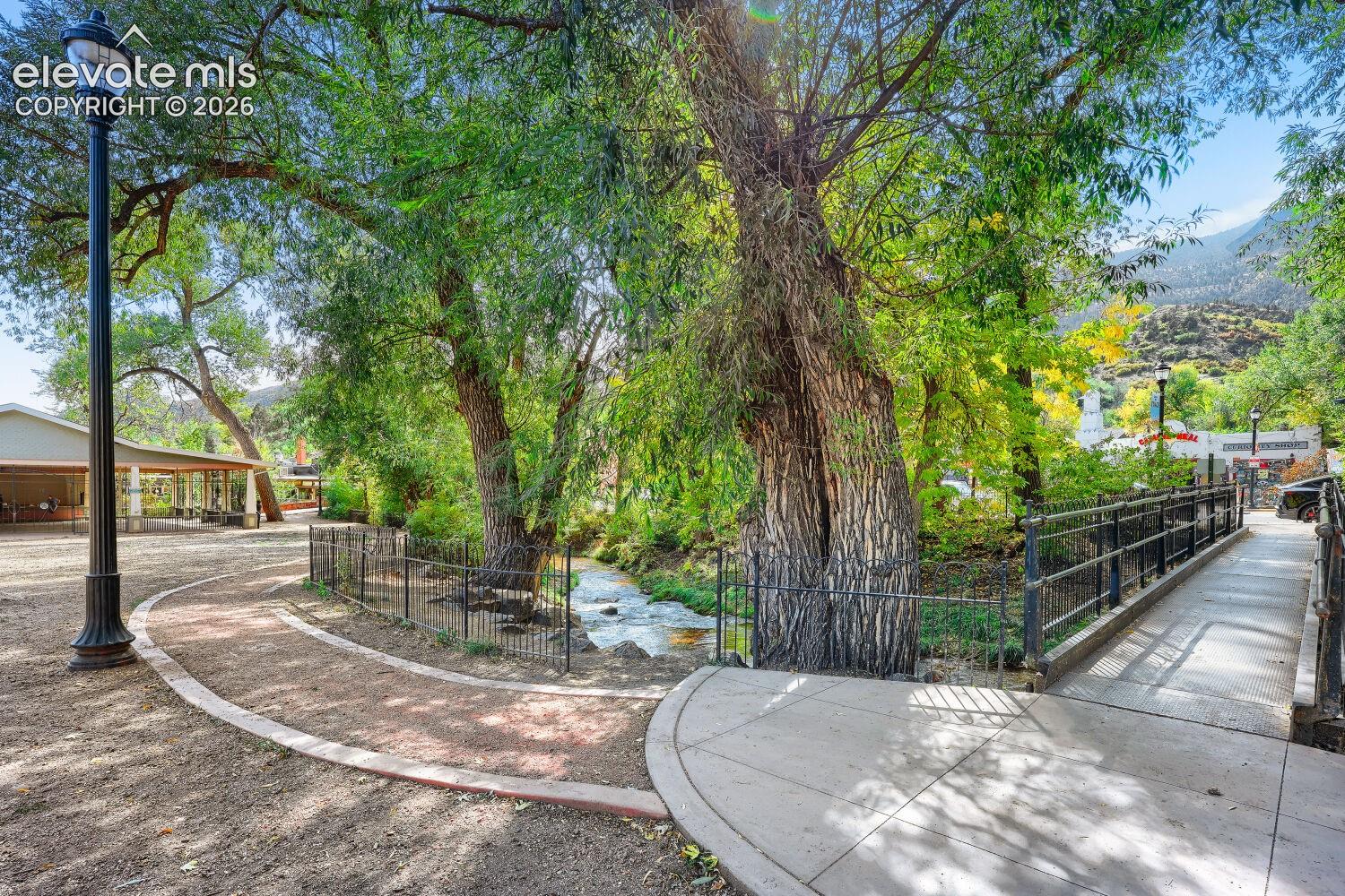 Paved pedestrian paths, a black metal bridge with railings, a flowing creek, and mature trees with lush foliage