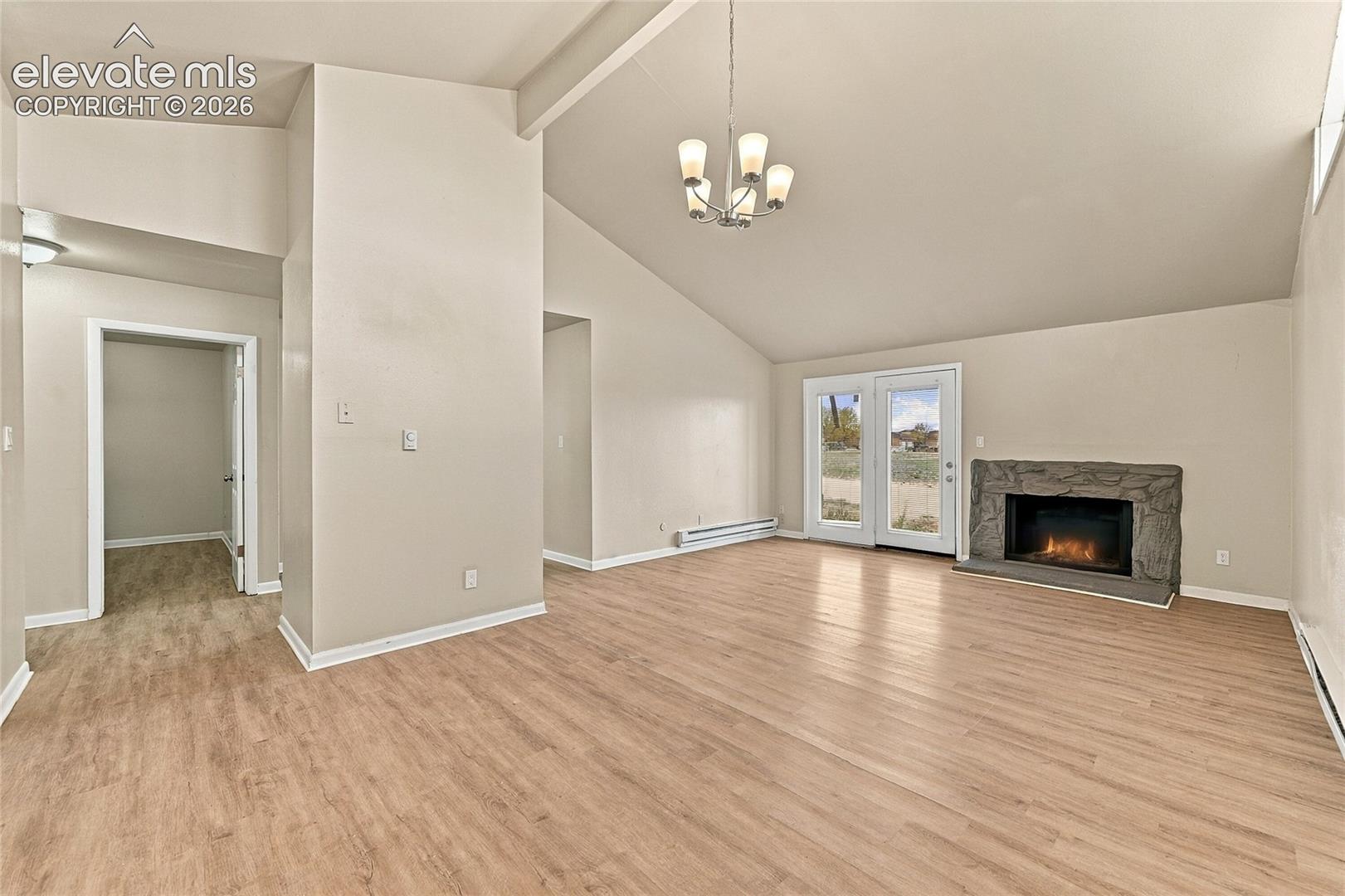 Vaulted ceiling, stone-surround fireplace, wood-finish flooring, multi-pane glass doors, and multi-arm chandelier