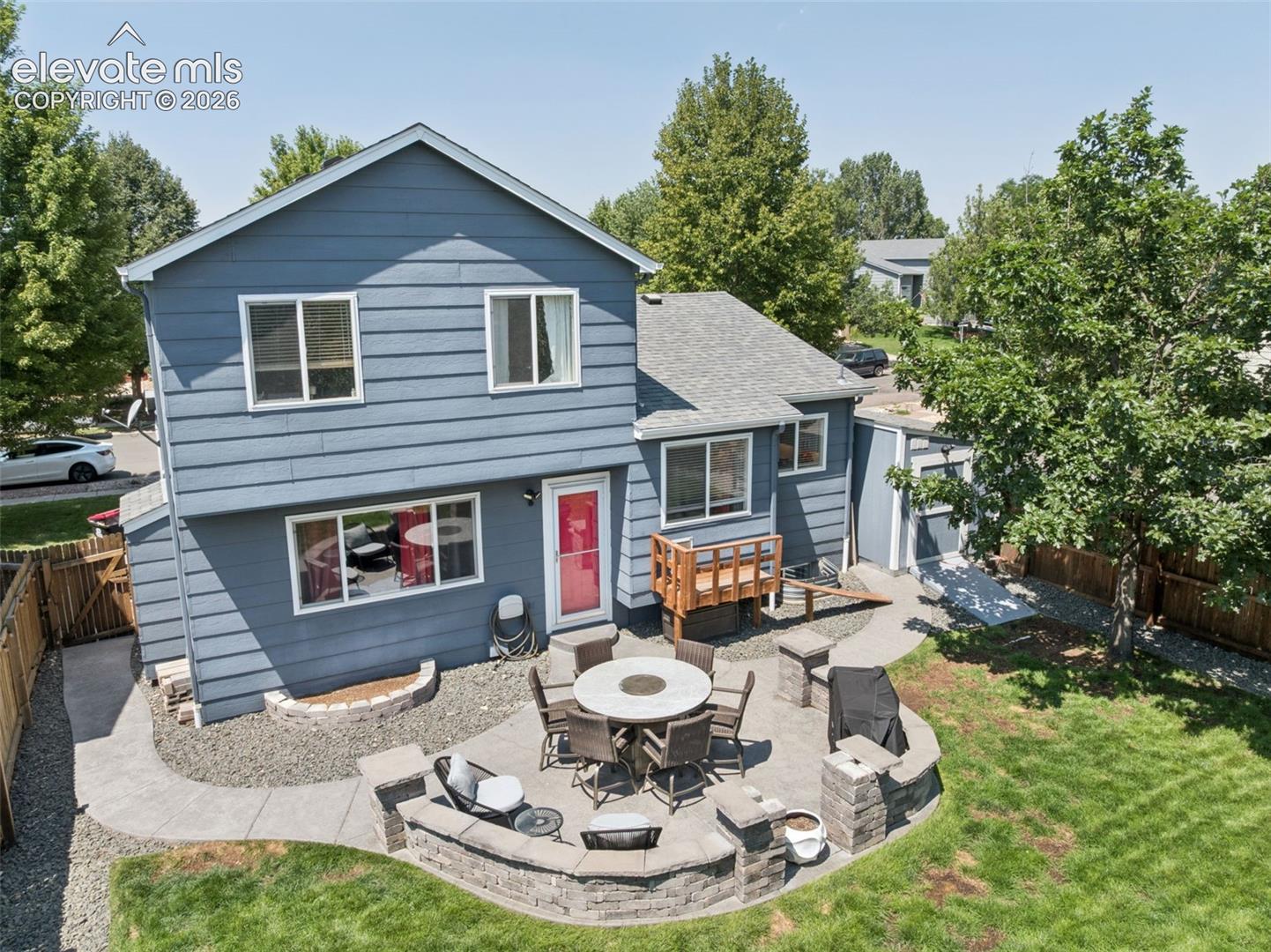 Two-story residence with blue siding, a multi-level patio with stone retaining walls, a wood-slat deck, and a fenced backyard