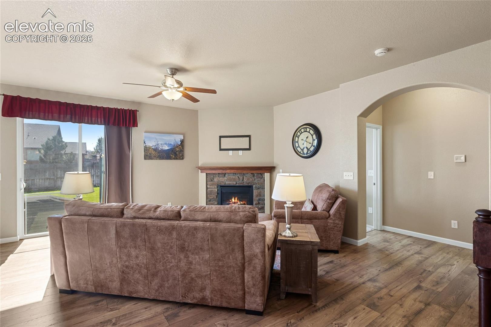 Living area featuring wood-finish flooring, a stone-surround fireplace with a wooden mantel, a ceiling fan, and sliding glass doors