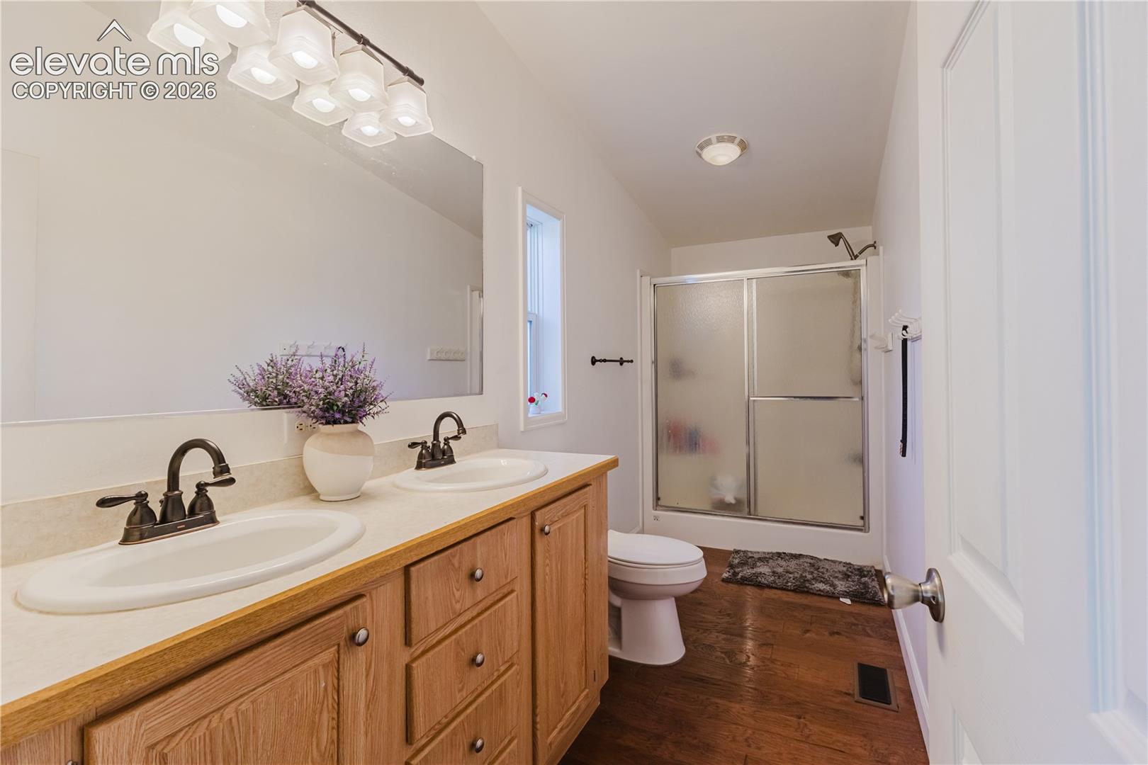 Double vanity with dual sinks and bronze-finish faucets, wood-finish cabinetry, full-width mirror, and a shower with sliding frosted glass doors