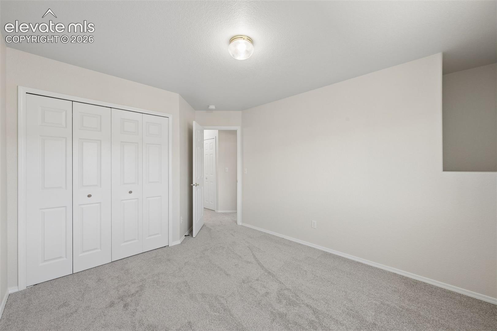 Neutral-toned interior space featuring light-colored wall-to-wall carpeting, white four-panel folding closet doors, and a flush-mount ceiling light fixture