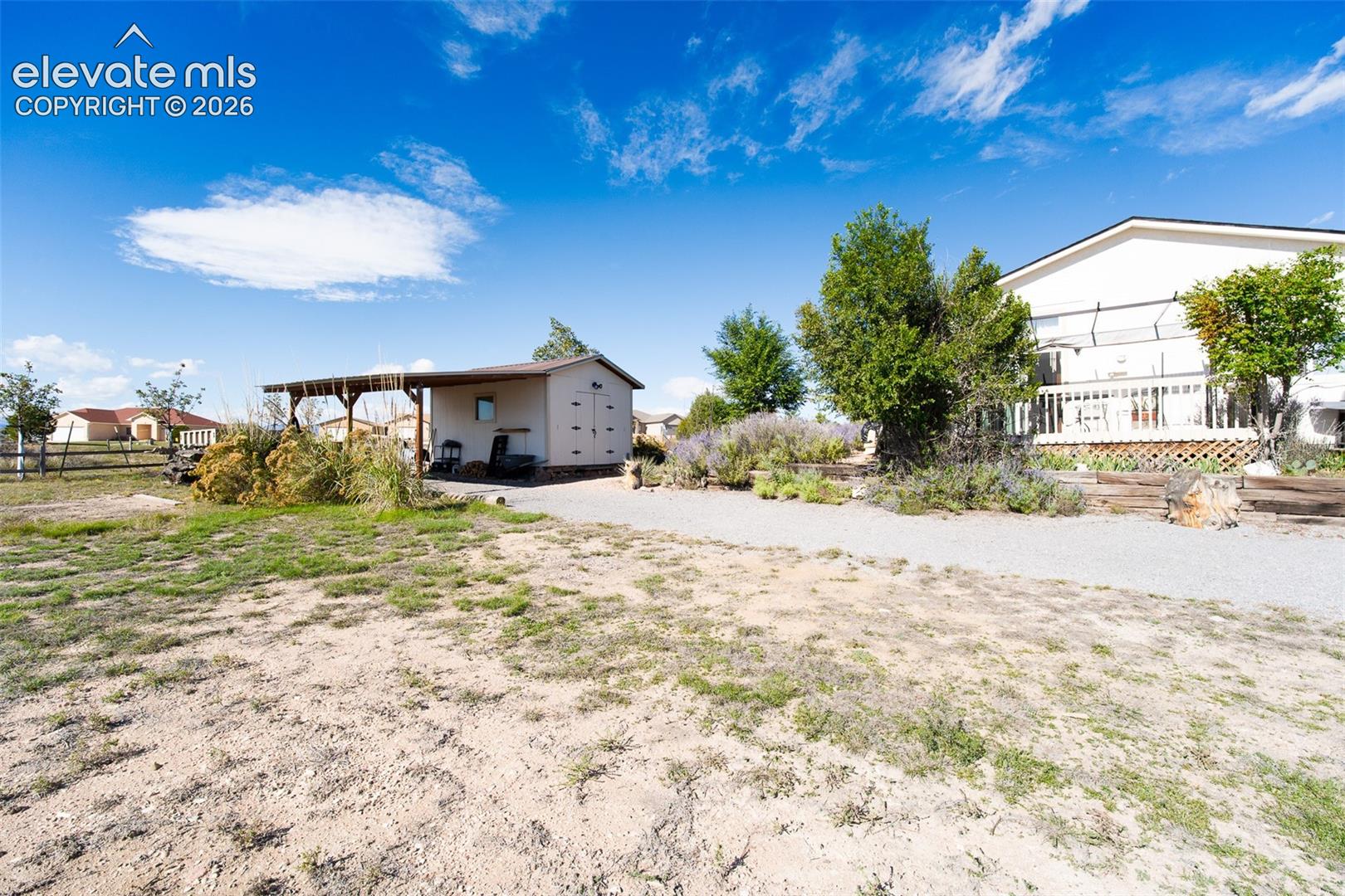 Expansive lot featuring a detached structure with an attached carport, gravel driveway, mature trees, and landscaping elements