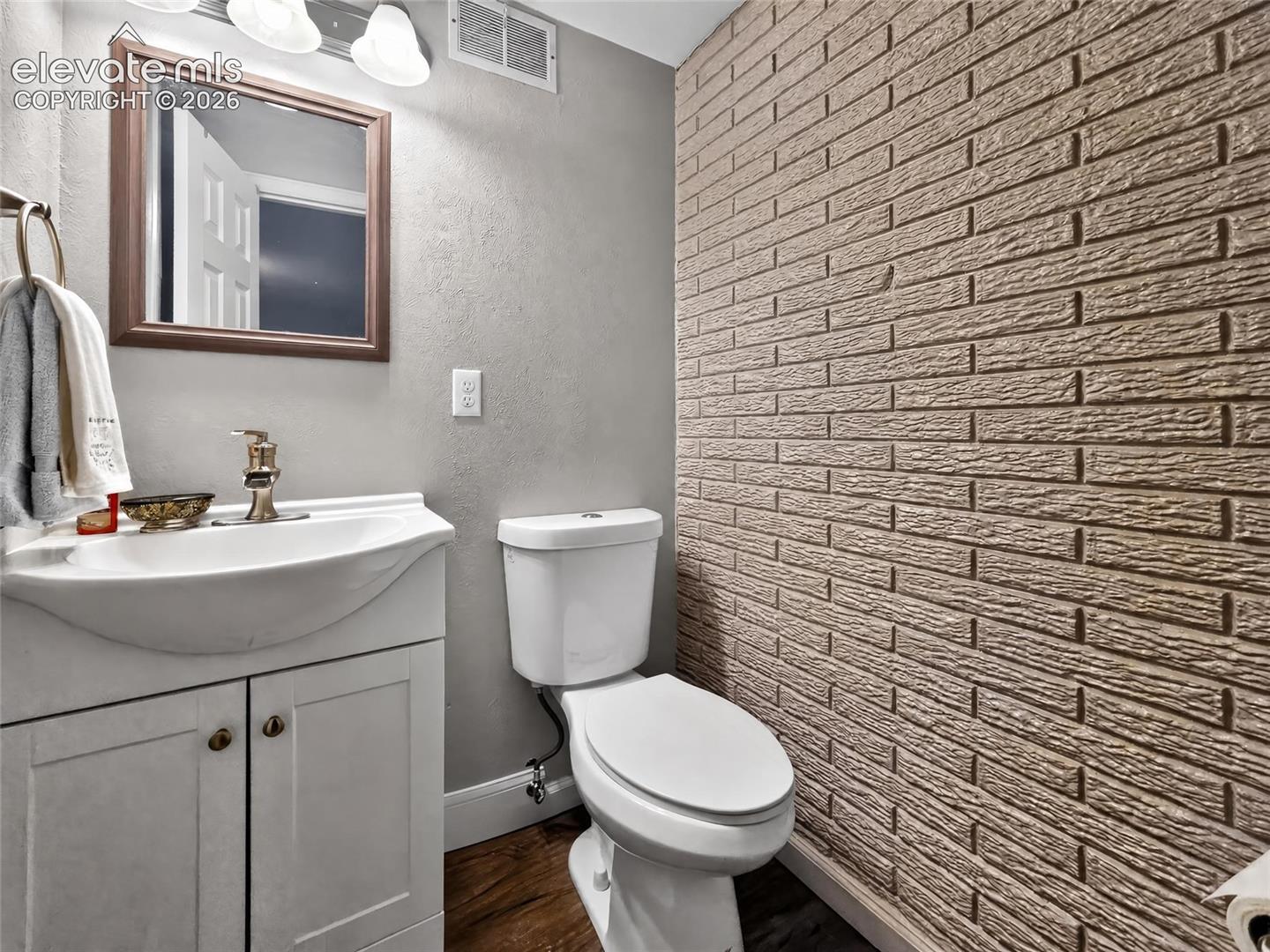 Textured brick-style accent wall, wood-finish flooring, white vanity with integrated sink, gold-toned faucet, and wall-mounted vanity lighting